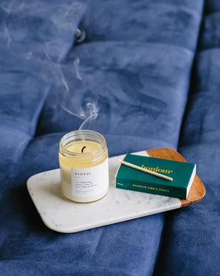 A lit candle with a wispy trail of smoke rises from an extinguished flame, placed on a white rectangular tray alongside a green matchbox with a burnt matchstick. All items rest on a soft, blue, quilted surface. The eco-friendly label reads "Santal Minimalist Candle by Brooklyn Candle Studio.