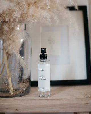 A minimalist scene featuring a clear glass spray bottle labeled "Santal Room Mist by Brooklyn Candle Studio," filled with essential oils, on a wooden surface. In the background, there's a large glass vase with pampas grass and framed artwork. The setting is soft and neutral-toned, evoking a calm and clean aesthetic.