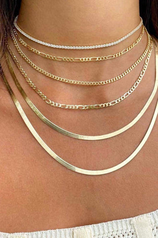 Close-up of a person wearing multiple layered gold necklaces of varying styles and lengths, including the dazzling Thin Tennis Choker by By Adina Eden adorned with CZ stones. The person is dressed in a white, textured, sleeveless top, with only part of their shoulders and upper chest area visible.