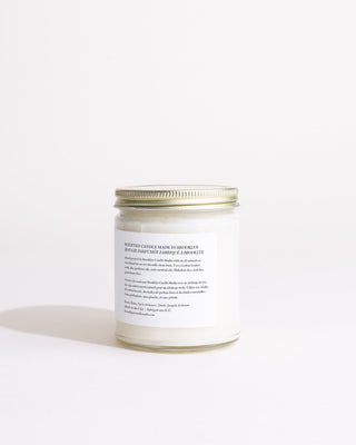 A clear glass jar with a metallic lid contains a white, eco-friendly scented candle made from soy wax. The label reads: "Sunday Morning Minimalist Candle by Brooklyn Candle Studio" and includes details about the candle's scent, burning instructions, and manufacturer. The background is plain white, emphasizing its cruelty-free nature.