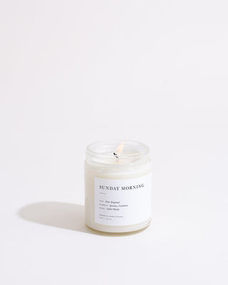 A lit Sunday Morning Minimalist Candle by Brooklyn Candle Studio, crafted from white soy wax and housed in a clear glass jar, stands against a plain white background. The label reads "SUNDAY MORNING" and features notes of pear, bergamot, jasmine, gardenia, amber woods, and musk. This eco-friendly and cruelty-free candle only reveals its delicate flame and a slight shadow.