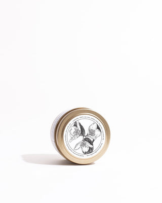 A small tin with a beige lid featuring an intricate black and white drawing of tulip flowers, identified as the Sunday Morning Gold Travel Candle by Brooklyn Candle Studio's eco-friendly line, lies on its side on a white surface, casting a soft shadow. The background is clean and white.