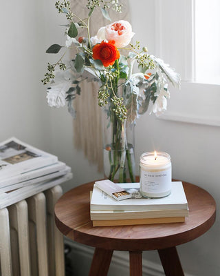 A small wooden table holds a vase of red and white flowers with lush green foliage. Beside the vase is a lit Sunday Morning Minimalist Candle by Brooklyn Candle Studio, offering an eco-friendly soy wax glow. A book, a stack of matches, and a couple of magazines are also neatly arranged on the table. The entire scene is elegantly illuminated by soft, natural light streaming in through the nearby window.