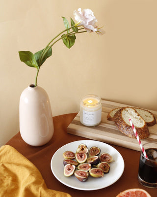 A ceramic vase with a single white flower, a lit Sweet Fig Minimalist Candle by Brooklyn Candle Studio, and a wooden cutting board with sliced bread are arranged neatly on a table. A plate of sliced figs, a partially visible glass with a red and white striped straw, and a mustard-colored cloth are also on the table.