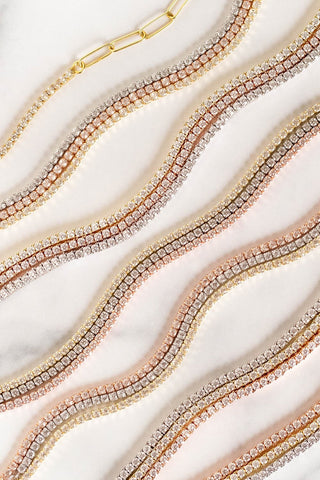 A collection of Thin Tennis Choker bracelets by By Adina Eden, available in various colors and featuring dazzling CZ stones arranged in wavy lines on a light marble surface. These luxurious bracelets come in gold-plated sterling silver and rose gold finishes, showcasing an elegant and refined appearance.