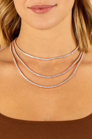 A woman with shoulder-length blonde hair wearing a brown ribbed top and the Three Prong Tennis Necklace by By Adina Eden, featuring three strands of sparkling cubic zirconia stones set in sterling silver. The photo, taken against a soft pink background, focuses on her neck adorned with this luxurious jewelry.